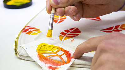 Eine Hand malt mit einem Pinsel bunte Blätter auf Stoff